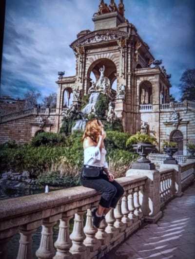 The image depicts a young woman sitting on a stone balustrade with a fountain in the background. The woman is wearing a white off-the-shoulder top and dark pants, with a black fanny pack around her waist. She is looking to her right and holding her hand up to her face. The fountain is elaborate, with multiple tiers and sculptures, surrounded by lush greenery. The sky is cloudy, with dramatic light and shadow patterns cast on the tiled ground. The scene appears to be in a park or public garden. The location is not Balakliya, Ukraine, but rather the Cascada Monumental in Barcelona, Spain.