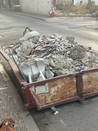 In La Línea de la Concepción, Spain, a construction debris skip is overflowing with broken tiles, concrete fragments, and what appear to be discarded plumbing fixtures, including two white ceramic sinks. The skip is red with rusty patches and sits on a paved street next to a sidewalk. In the foreground, the back of a brown dog wearing a black collar is visible, suggesting a person is present but out of frame. The background shows a street with a pedestrian crossing, a building with a tiled facade, and a glimpse of vegetation. The scene appears to be captured during daylight.