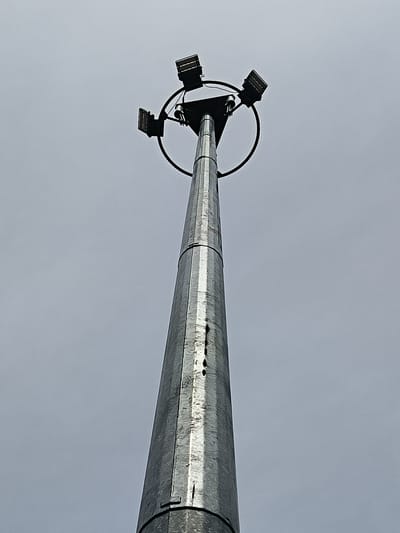 A tall, metallic pole with four floodlights at its apex stands against a pale, overcast sky. The pole tapers as it rises, its surface showing some wear and tear. The floodlights are angled outwards, suggesting they are positioned to illuminate a large area. The scene is devoid of people or any other visible objects.