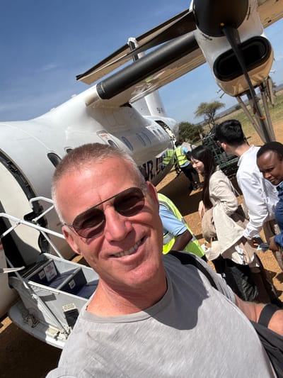 A man wearing sunglasses smiles in a selfie with an airplane in the background. Passengers board. Two men in business attire and a woman wearing a neon yellow vest are visible near the plane. The plane is white with black accents. The sky is clear and blue. Boxes labeled "for use only at a remote airport" are visible on steps near the plane's entrance. The man's face is in focus.