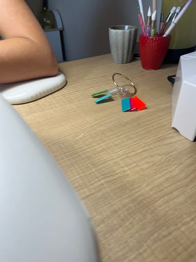 The image shows a close-up view of a wooden desk surface. Various objects are arranged on the desk, including a red ribbed cup filled with brushes, a metallic gray cup, and a keyring with colorful rectangular charms. In the background, a white object, possibly a box or a piece of equipment, is partially visible. A person's leg and knee are also in the foreground, positioned on the right side of the frame. The scene appears to be indoors, with the lighting suggesting it could be daytime. The overall impression is one of a workspace or a craft area. There is no visible text in the image.