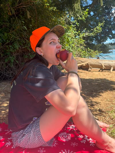 A person wearing a black t-shirt, patterned shorts, and an orange baseball cap sits on a red blanket outdoors. They are holding a peach to their open mouth, about to take a bite. The person is sitting on a sandy area with foliage and trees in the background. A body of water and a large fallen tree are visible in the distance. The scene appears to be in a natural setting, possibly a park or beach area.