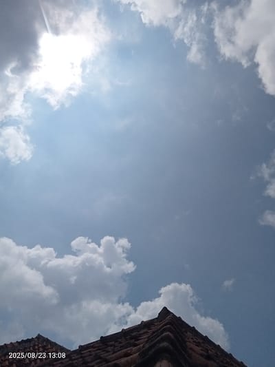 The image depicts a bright, partly cloudy sky observed from a low angle, with a section of a tiled roof visible in the lower foreground. The sun is prominently featured in the upper left quadrant, appearing as a bright, overexposed light source partially obscured by white and grey clouds. A faint, linear contrail is visible extending diagonally from the upper left. The sky transitions from a lighter blue near the sun to a deeper blue in the upper right. White, fluffy cumulus clouds are scattered across the frame, with a larger cluster near the bottom center and right. The lower portion of the image shows the peak and right slope of a gabled roof, constructed with reddish-brown or terracotta-colored tiles, exhibiting shadows and textural details. No people, actions, or interactions are visible. A digital timestamp "2025/08/23 13:08" is present in the bottom left corner. The scene is located in Palasari, Indonesia.