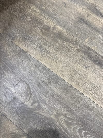 The image shows a close-up, angled view of a textured wooden floor, likely laminate or engineered wood, with a grayish-brown hue and prominent grain patterns. The planks are laid diagonally across the frame, with visible seams between them. There are subtle variations in color and texture, with some areas appearing lighter and smoother, and others darker and rougher, suggesting natural wood grain or wear. Some faint smudges or dust specks are visible on the surface. The lighting appears to be natural and diffused, casting a soft glow on the floor. The scene does not depict any specific Izium landmarks or recognizable urban elements.