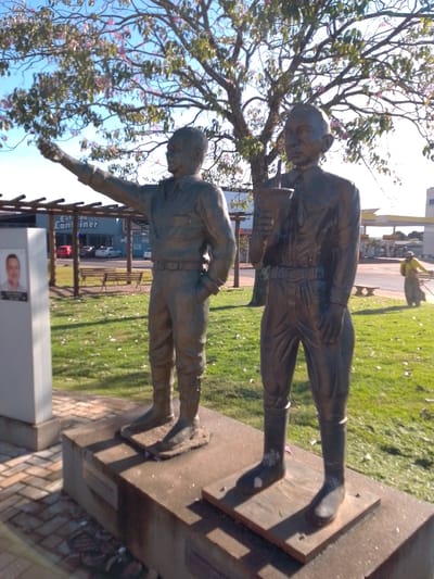 The image depicts a street scene in São Gabriel do Oeste, Brazil, with two bronze statues of men in uniform standing on a raised concrete platform. The statue on the left is pointing forward with its left arm, while the statue on the right is holding a cup-like object in its right hand. Both statues are dressed in military-style uniforms with boots. Behind the statues, a tree with pink flowers is visible. In the background, there is a building with text that appears to read "Estado Container," and further back, a gas station. A person in a yellow shirt and dark pants is seen walking in the distance, possibly holding a broom. The ground is covered with grass and paved areas, and there are several benches and a pergola-like structure. The sky is clear and blue.