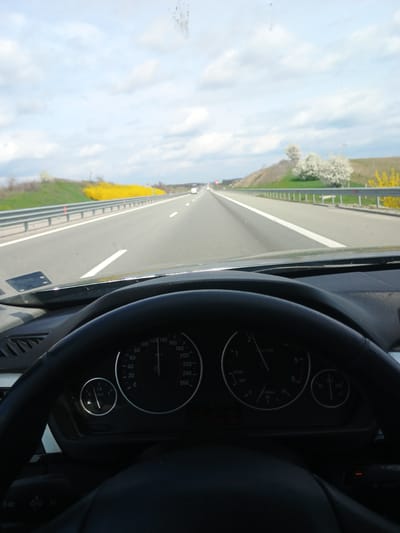 The image shows the view from inside a car driving on a highway. The steering wheel and dashboard are visible in the foreground. The speedometer reads about 140 km/h, and the tachometer indicates around 3000 RPM. Through the windshield, a straight highway stretches into the distance. The landscape beside the road features a line of bright yellow flowers/bushes and some green grass, with a light blue sky overhead. The location is not discernible as being in Malko Tranovo, Bulgaria from the image alone.