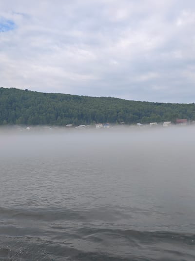 The image depicts a foggy morning scene on the Yenisei River in Atamanovo, Russia. A dense layer of fog hovers over the water, obscuring the lower portions of the houses that line the shore. The houses appear to be small, rural dwellings with visible roofs and windows. Behind the houses, a dense, green forest covers a hillside that slopes upwards to the horizon. The sky above is overcast with pale, diffused light. The water in the foreground is rippled, with some white foam visible at the bottom left, suggesting movement.