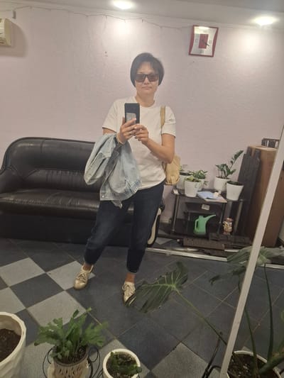 A woman stands in front of a mirror, taking a selfie. She is wearing a white t-shirt, dark jeans, and light-colored sneakers. She is holding a light blue denim jacket over her left arm and a mobile phone in her right hand, which she is using to take the photo. She is also wearing sunglasses. The room appears to be a living space or a small shop, with a black leather sofa to her left. In the background, there is a clock on the wall, a shelf with several potted plants, and various objects including a green watering can and a decorative item. The floor is tiled in a checkerboard pattern of dark grey and lighter grey squares. The overall lighting suggests an indoor setting. There is no specific indication of the city of Svobodny, Russia, or any identifying landmarks.