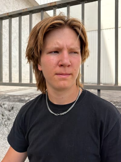 A young person with light brown hair and freckles is pictured from the chest up, looking to the right. They are wearing a black t-shirt and a silver chain necklace. The background features a light-colored building with vertical panels behind a metal railing. The person appears to be outdoors, possibly in an urban setting like Caluire-et-Cuire, France, given the architectural elements. The lighting suggests it is daytime.