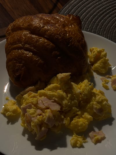 A close-up overhead shot shows a breakfast plate with a croissant and scrambled eggs. The croissant is on the left side of the plate, appearing golden brown and flaky. To the right of the croissant are scrambled eggs, mixed with small pieces of ham. The eggs are a bright yellow, and some pieces of ham are visible with pinkish-white coloration. The plate is white and sits on a dark wooden surface, possibly a table. A patterned placemat is visible on the left side of the frame, behind the plate. The lighting suggests it may be morning or evening, with a strong light source casting shadows on the food and plate. The scene depicts a simple breakfast. No text is visible in the image. The location context is Słupsk, Poland.