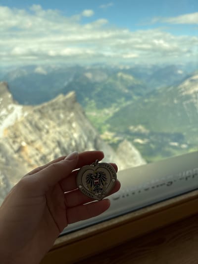 A hand is holding a heart-shaped keychain with the Austrian coat of arms and the text "ÖSTERREICH" and "AUSTRIA". The keychain is silver and has small rhinestones. The background is a panoramic view of the Alps, with blurred mountains and a cloudy sky. The scene is likely from the Zugspitze, which is the highest peak in Germany and borders Austria, and is a popular tourist destination accessible from Ehrwald, Austria.