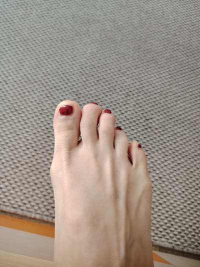 A close-up, top-down shot shows a single bare foot positioned on a textured, grey surface. The foot has five toes, all painted with dark red nail polish. The skin is fair, and the foot appears clean. The background is a uniform grey fabric with a small, repeating pattern of raised dots. The lighting is soft, creating subtle shadows and highlighting the contours of the foot. There is no indication of location in the city of Putilkovo, Russia, nor any other environmental context beyond the immediate surface the foot rests upon.