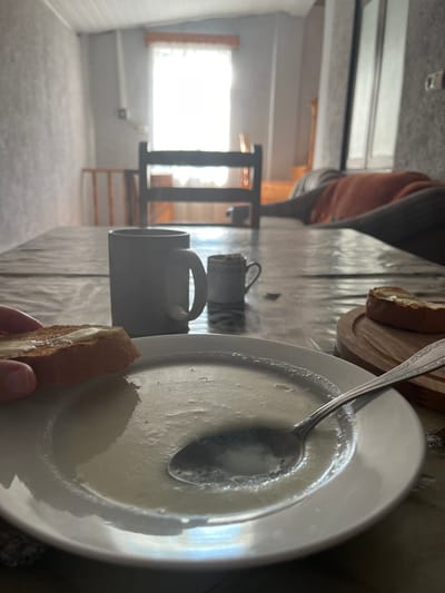 A close-up, eye-level shot shows a person holding a piece of buttered bread over a white plate of pale soup with a spoon in it. The bread is toasted and has melted butter. Behind the soup, two mugs are visible on a table covered with a patterned plastic cloth. The background is blurry, showing a window with white curtains, a wooden chair, and a sofa with an orange blanket. The scene appears to be indoors, possibly a dining area in a home within ღრმაღელის თემი, Georgia, characterized by its simple, rustic interior.