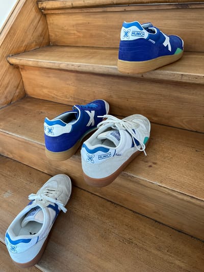 The image depicts an indoor wooden staircase in Foetz, Luxembourg, featuring four Munich Gresca athletic shoes distributed across three steps. No individuals are present.
On the uppermost visible step rests a single blue Munich Gresca sneaker, oriented with its heel partially facing away from the observer. This shoe features a blue suede and textile upper, a white "X" logo, "MUNICH" branding on the heel counter, white laces, and a light brown gum sole.
The middle step holds two sneakers. A second blue Munich Gresca sneaker, consistent with the first, is positioned with its toe pointed towards the lower left. Adjacent to it, a white Munich Gresca sneaker is placed with its toe pointed towards the lower right. This white shoe exhibits an off-white suede and textile upper, a blue "X" logo, blue "MUNICH" branding with "88" detail on the heel, white laces, and a light brown gum sole.
On the lowest visible step, a second white Munich Gresca sneaker, matching the one on the middle step, is positioned with its toe pointed towards the lower left.
The staircase is constructed from light-colored wood, displaying visible grain and minor surface wear on the treads and risers. The scene is evenly illuminated by ambient light.