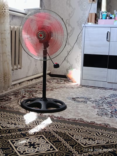 A floor fan with a red-tinted head and a black stand is positioned on a rug. The fan's blades are blurred, indicating it is in motion. To the left of the fan, a portion of a window with light-colored curtains and a radiator are visible. In the background, there is a wall with patterned wallpaper and a white cabinet with dark accents. A plastic bag with colored contents is on the floor in front of the cabinet. The timestamp "17.06.2026 09:42" is displayed in the bottom right corner of the image. The scene appears to be an indoor setting, likely a residential room.