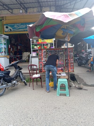 The image shows a street scene in Keudematangglumpangdua, Indonesia. A man wearing a black shirt and blue jeans is standing at a street vendor stall displaying eyeglasses and watches. A multi-colored sun umbrella shades the stall. The stall is set up near a shop with a sign reading "Warkop Sama" and a smaller text in Indonesian below. A motorcycle is parked to the left of the stall, and another is further down the street, along with a few other people. A red plastic chair is near the motorcycle, and a green plastic stool is next to the man at the stall. The street is paved.