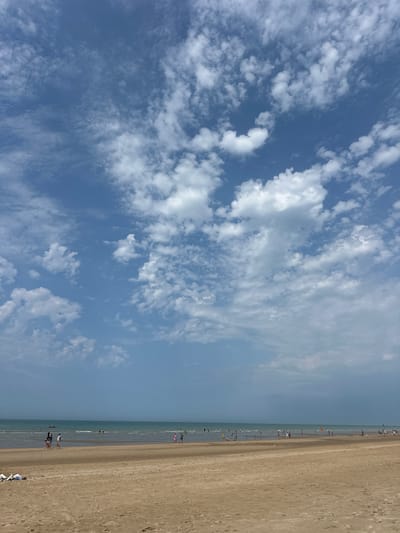 The image depicts a wide, sandy beach in Koksijde, Belgium. The foreground is dominated by the pale, granular sand, with a few scattered items of clothing lying on the left side. Further back, the sea stretches to the horizon, its surface reflecting the blue sky. The water is calm with gentle waves breaking on the shore.

A diverse group of people are present on the beach and in the shallow water. Some are walking along the shoreline, while others are wading or standing in the sea. There are also indications of people further out in the water, possibly swimming or engaged in water activities. A small red boat is visible on the water in the distance to the left.

The sky is a prominent feature, filled with a dramatic display of white and grey clouds against a clear blue backdrop. The clouds vary in texture, from wispy, scattered formations to larger, more defined cumulus shapes. The lighting suggests it is daytime, likely midday or early afternoon, with the sun illuminating the scene. The overall atmosphere is one of a pleasant, active beach day in a coastal environment.