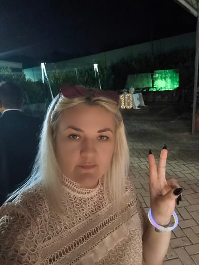 A medium shot captures a fair-skinned woman with blonde hair and dark eyebrows, wearing a light-colored lace top. She is making a peace sign with her right hand, which has black nail polish and a glowing wristband. In the background, a man in a suit is visible on the left. The scene appears to be an outdoor event at night, possibly a celebration, with illuminated decorations that resemble the number "18". The ground is paved with bricks, and there is greenery and artificial lighting in the background. The location is identified as Vinchiaturo, Italy.