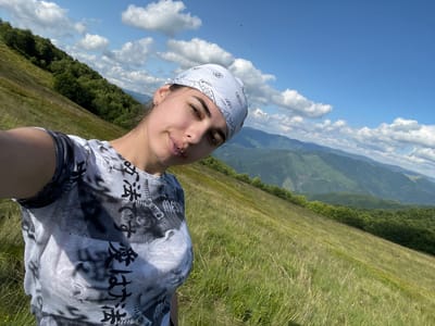 This is a vertical, low-angle selfie taken outdoors in a mountainous region, likely in Ust'-Chorna, Ukraine, under a clear blue sky with scattered white clouds. The scene is dominated by a vast expanse of green grass, indicative of a summer meadow, with distant rolling hills and a forest line visible in the background. The time appears to be daytime, given the bright sunlight casting shadows.

The main subject is a young woman with fair skin and dark hair, wearing a white bandana with a black paisley pattern and a grey t-shirt featuring black text and imagery. The text on her shirt appears to be in Japanese characters. She is looking directly at the camera with a slightly smirking expression, suggesting a relaxed and playful mood. She has a small diamond earring in her visible ear. The photo captures a sense of freedom and enjoyment of nature.