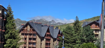 The image depicts a mountain resort setting in Sallent de Gállego, Spain, on a clear, sunny day with a bright blue sky. In the foreground, several multi-story buildings with stone facades and dark, pitched roofs dominate the scene. These buildings feature numerous balconies with wooden railings. Tall evergreen trees, likely pine or fir, are interspersed between the buildings, adding to the alpine atmosphere.

In the background, a range of majestic mountains rises, with some of their peaks partially obscured by wispy white clouds. The lower slopes of the mountains are covered in green vegetation, indicating a summer or early autumn season. The architecture of the buildings suggests a style common in ski resorts, designed to blend with the natural mountainous environment. The overall impression is one of a peaceful, scenic mountain town. There are no people visible in the image, and no actions or interactions are depicted.
