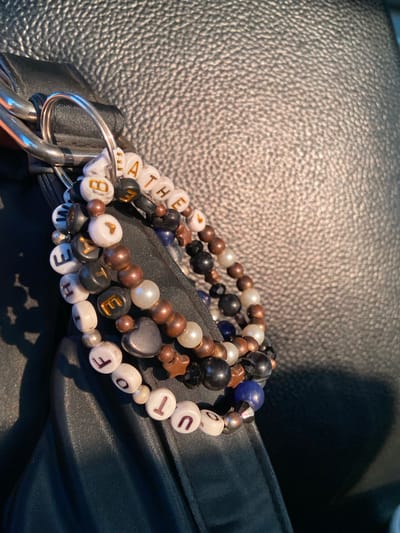 The image shows a close-up of beaded bracelets and charms attached to a keychain ring. The bracelets are made of various beads, including white letter beads, dark colored beads, and metallic copper-colored beads. Some of the letter beads spell out words. There are also some decorative charms, such as a dark heart-shaped charm and star-shaped charms. The keychain ring is silver-colored. The bracelets and keychain are resting on a black, textured surface, likely leather or faux leather. In the background, a dark, slightly shiny fabric is visible. The lighting suggests it is daytime, with shadows cast from the objects. The image does not provide any information about the city of Jekulo, Indonesia, or any people or actions.