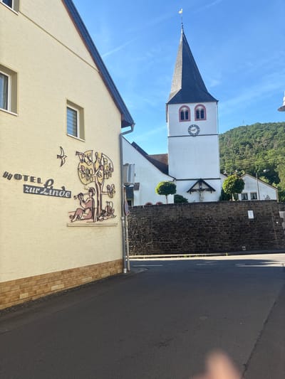 This is a medium-shot, eye-level, outdoor photograph captured on a sunny day in Schuld, Germany. The scene depicts a street with a building on the left and a church steeple in the background.

On the left, a cream-colored building with a brown brick base has a decorative mural on its wall. The mural features the words "HOTEL zur Linde" in dark lettering, with a stylized tree and a person playing a flute beneath it. A bird is flying above the tree. The building has at least two windows with white frames.

To the right of the building, a white church with a dark steeple rises into the blue sky. The steeple has two arched windows and a clock face. A cross with a statue of Jesus is mounted above the entrance to the church. The church is surrounded by a low stone wall. Behind the church, a lush green hillside is visible, dotted with trees. The street in the foreground is asphalt, with a curb and some patches of grass.

The lighting suggests it is daytime, with strong shadows cast by the building and the church. The overall impression is one of a tranquil village setting.