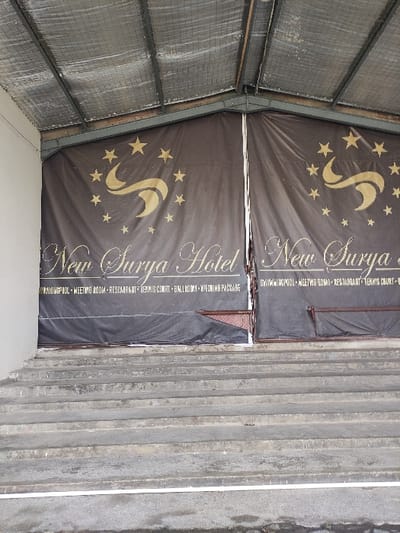 Two banners with the text "New Surya" and "New Surya Hotel" are mounted on the side of a building. The banners are made of black fabric with gold lettering and stars. The banners are hung vertically. The building is made of concrete and has a grey, rough exterior. The banners are positioned at an angle to each other. The scene is viewed from a low angle, looking up at the banners. The location is Gambiran, Indonesia, at coordinates -8.434, 114.18.