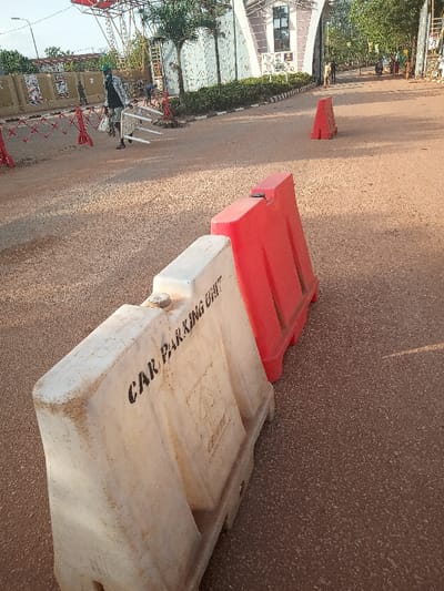 The image shows two traffic barriers in a paved road in front of a building in Otta, Nigeria. One barrier is white with the words "CAR PARKING UNIT" printed on it, and the other is red. The white barrier is in the foreground, and the red barrier is behind it, partially obscured by the white barrier. The red barrier is lying on the ground, and the white barrier is standing upright. The road is empty except for the barriers. The pavement is dusty and brown.  The image is taken from a low angle, looking up at the barriers. There are no people or vehicles visible in the image. The location is in Otta, Nigeria, at the coordinates 6.682 latitude, 3.168 longitude.
