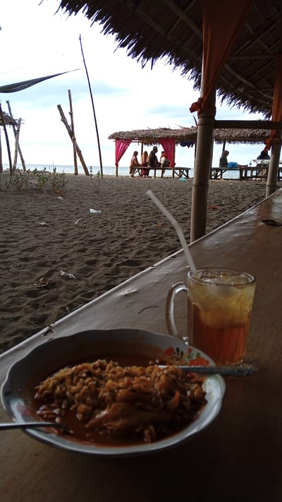 A beachside scene in Pasi ie Leubeue, Indonesia, shows a thatched-roof structure with orange fabric drapes, a common sight at local eateries. In the foreground, a meal of what appears to be noodles in sauce is served in a white bowl with a floral pattern. Beside it, a glass of iced tea with a straw sits on a wooden table. The beach, covered in dark sand, has scattered debris. In the mid-ground, several people are seated at tables under similar thatched structures, enjoying the seaside view. The ocean is visible in the background under a partly cloudy sky, with some wooden poles and a dark net structure suggesting a simple beach setup.