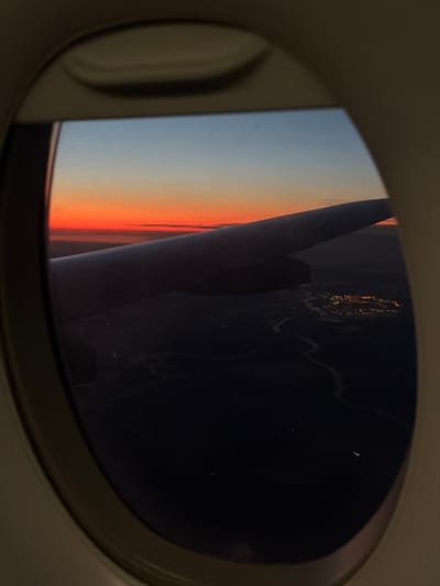 The image is taken from an airplane window, looking out at a sunset over a landscape. The sky transitions from a deep blue at the top to a vibrant orange and red near the horizon. A portion of the airplane's wing is visible in the foreground, extending diagonally across the frame. Below the wing, a dark landscape is illuminated by a cluster of city lights, suggesting a populated area. A winding river or body of water is visible near the city lights. The specific location in the city of Шемякино, Russia cannot be determined from the visual information provided in the image.