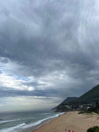 A beach scene under a cloudy sky. A mountain range runs parallel to the shoreline. Waves roll onto the sandy beach. A red and white object, potentially a vehicle, sits on the sand. The cloudy sky dominates the upper portion of the image, with hints of blue breaking through. Buildings are nestled into the vegetation-covered mountain.