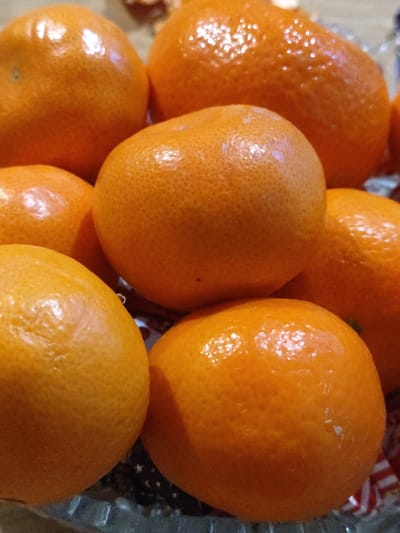 The image shows a close-up of a pile of tangerines in a glass bowl. The tangerines are bright orange, glossy, and display a textured surface. The bowl appears to have a red and white striped pattern at the bottom. The location in the city of Yerzhovo, Moldova is not discernible from the image.