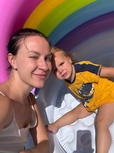 In this image, two individuals, a woman and a young child, are depicted in what appears to be an inflatable pool with a rainbow-colored interior. The woman is in the foreground, her face close to the camera, with her eyes partially closed and a gentle smile. Her dark hair is wet and slicked back. She is wearing a white camisole with lace trim.
The child, positioned above and behind the woman, is also upside down relative to the viewer. The child is wearing a yellow t-shirt with a dark blue collar and sleeve trim, featuring a graphic of a stylized creature. The child's face is visible, and they appear to be looking towards the camera with a slight smile.
The setting is an inflatable pool, with the rainbow colors of the pool walls forming a curved backdrop. The bottom of the pool is a white, textured surface, likely also inflatable. Sunlight is casting distinct shadows on the white surface, suggesting it is daytime and the sun is bright. The overall mood is relaxed and playful, indicative of a leisurely activity like playing in a pool. No text is visible in the image. The location context provided places the scene in the Astrashitskagaradokski rural council, Belarus.