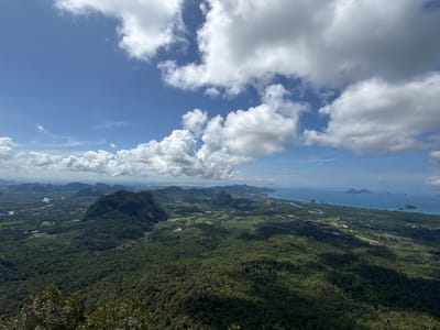 The image presents a wide-angle aerial view overlooking a lush, green mountainous landscape. The foreground is dominated by the dense canopy of a forest, with some vegetation visible at the very bottom edge, suggesting the vantage point is from a high elevation. In the midground, rolling hills covered in trees stretch out, interspersed with scattered buildings and some cleared areas that might be agricultural land or small settlements.

Further into the distance, the landscape transitions to a more varied terrain with several prominent, steep, jungle-covered limestone karsts. Beyond these formations, a coastal area is visible, characterized by a sandy shoreline and the bright blue expanse of the Andaman Sea. Several small islands are dotted across the water, and the coastline extends into the horizon, showing signs of development with buildings clustered along the shore.

The sky above is a vibrant blue, punctuated by large, fluffy white cumulus clouds, casting shadows on the terrain below. The lighting suggests it is daytime, likely late morning or early afternoon, with the sun intermittently breaking through the clouds.

While the location is stated to be Ban Din Daeng Nui, Thailand, the image itself does not contain any specific landmarks or text that definitively identify this precise location. However, the scenery aligns with the characteristic karst topography and coastal environment of southern Thailand. There are no people, animals, or specific actions depicted in the image. The scene is a natural landscape captured from a high vantage point, emphasizing the scale and beauty of the environment.