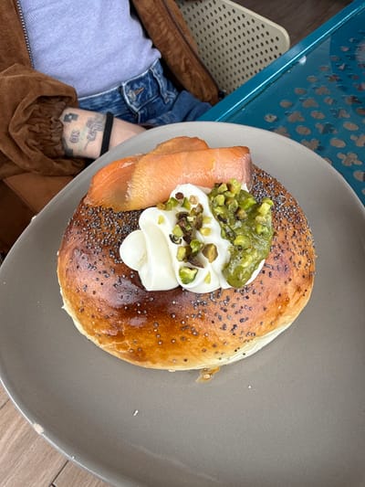 An overhead view captures a meal served at an outdoor establishment in Playa Blanca, Spain. The central subject is a large, golden-brown, poppy-seed-topped pastry, resembling a brioche or a bagel, presented on a round, matte grey plate. It is garnished with several folded slices of smoked salmon, a dollop of white cream, and a portion of green, chunky pistachio pesto, all topped with chopped pistachios. To the upper left, the torso and left arm of a person are visible. This individual wears a light-colored top, a brown zippered jacket, and blue denim jeans. The visible forearm displays tattoos, including the letters "OK" and another symbol, with a black elastic band around the wrist. The plate rests on a light-colored floor or surface. To the right of the plate, a turquoise perforated metal table with decorative cutouts is visible, alongside a light-colored perforated chair in the background, suggesting an outdoor cafe or restaurant setting.