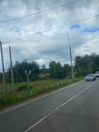 The image captures a roadside scene on a cloudy day, likely in or near Почтовик Снт, Russia. A paved road with a white line marking runs from the foreground into the distance. On the left side of the road, tall power poles and overhead wires are visible, along with a railway crossing signal with two round lights. Beyond the road and a grassy verge lined with yellow markers, there is a dense line of trees and foliage. Two cars are traveling on the road, one silver sedan in the middle lane and a white SUV behind it. The sky is overcast with a mix of dark and lighter clouds.