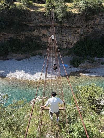 A high-angle, long shot captures three individuals traversing a narrow, rustic suspension bridge spanning a deep gorge over a clear, turquoise-colored river. The bridge, constructed from rusted metal grating forming the walkway and thick steel cables for support, extends from the foreground into the distance.

In the immediate foreground, a male figure, viewed from behind, stands with his hands on the bridge's side railings. He wears a light-colored short-sleeved shirt, dark shorts, and white sneakers. Further along the bridge, a second male figure, also seen from behind, walks in a white t-shirt, dark shorts, and white sneakers. A dark object, possibly a small bag, rests on the bridge near him. At the far end of the bridge, a third figure, in dark clothing, approaches the opposite bank.

The river below features wide, white pebble and gravel banks, with a visible blue tarp or fabric item on the far bank. Steep, rocky cliffs rise on both sides of the river, densely covered with green trees, shrubs, and foliage. Sunlight illuminates the scene, creating distinct shadows on the riverbanks and bright glints on the water. The environment suggests a remote, natural landscape in the city of Unknown, Montenegro.