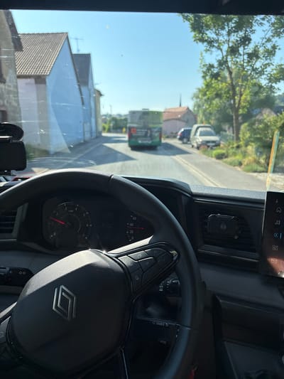 The image is taken from the interior of a vehicle, looking forward through the windshield. A steering wheel with the Renault logo is prominently displayed in the foreground. The dashboard includes a speedometer, tachometer, and other controls. The vehicle appears to be driving on a road in a village, with houses and trees lining the street. In the distance, a green bus is visible. The sky is clear and blue. The scene is consistent with a sunny day. The overall impression is that of a vehicle traveling through a European village.