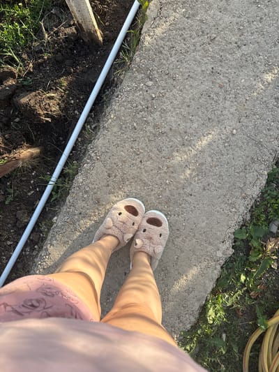 This is a top-down, close-up shot of a person's legs and feet, wearing fluffy light brown slippers with bear-like features and pink shorts. The person is standing on a concrete path. To the left of the path, there is disturbed dark soil with some green grass and a metal object. A white pipe runs along the edge of the path and the soil. To the right of the path, there is more green grass and a yellow hose coiled on the ground. The scene appears to be outdoors, in what might be a garden or yard area, possibly in the city of Sovkhoza "Razdol'e", Russia. The lighting suggests it is daytime, with sunlight casting shadows on the concrete.