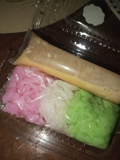 The image depicts a clear plastic food container with a lid, resting on a dark, possibly wooden, surface in Doramamidi, India. Inside the container, there are four individually packaged food items. Three smaller clear plastic bags contain portions of noodle-like or gelatinous strands, colored pink, white, and green, arranged side-by-side. A larger clear plastic bag contains a yellowish-brown liquid. No people are visible.