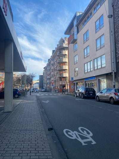 The image is taken from a low angle looking up at a building facade at the corner of Rue de la Victoire and Avenue de la Victoire in Etterbeek, Belgium. The building is a pale beige color with large rectangular windows. There is a blue sign on the building that reads "MINI MAX". The street is paved with gray cobblestones and has a bike lane marked with white paint. There are several parked cars on the right side of the street. The sky is a bright blue with white clouds. There are no people visible in the image. The scene is deserted and quiet. The image is taken from a street level viewpoint. The composition is angled, with the building in the foreground and the street in the background. The lighting is bright and sunny. The overall tone of the image is calm and peaceful.