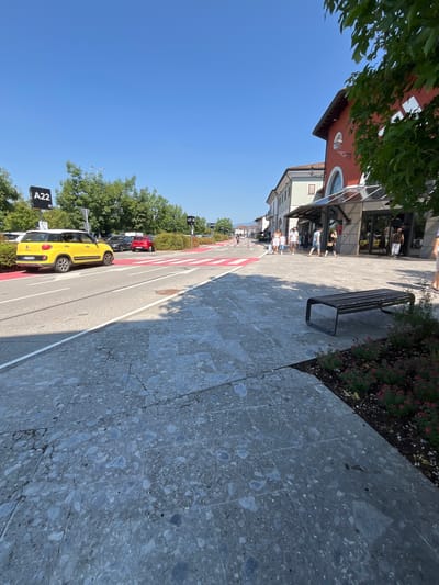 This image depicts a sunny day in Rodengo, Italy. A bright yellow Fiat 500L is parked on the left side of the road, next to a red-lined pedestrian crossing. Further down the road, other cars and people can be seen walking in front of shops. A modern, dark-colored bench sits on a stone-tiled sidewalk to the right, adjacent to a flower bed with small pink flowers. The sky is clear blue, and trees line the street.