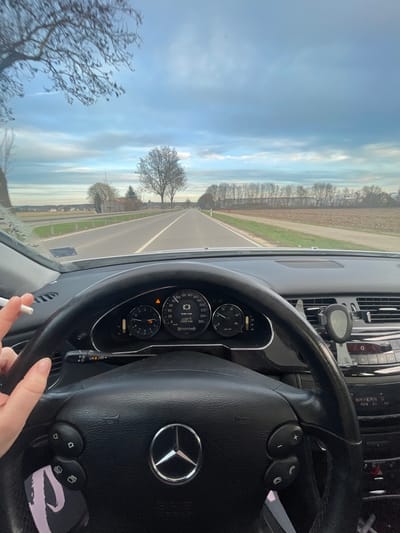 A first-person perspective from the driver's seat of a Mercedes-Benz vehicle, looking forward onto a two-lane asphalt road in a rural area near Weißenhorn, Germany. The driver's right hand, with short, manicured nails, holds a lit cigarette with ash visible at its tip, positioned just above the black leather multi-function steering wheel. The steering wheel prominently displays the Mercedes-Benz star logo. The instrument cluster shows a 'BRAKE WEAR' warning, an outside temperature of 13.0°C, and an odometer reading of 200,438 km. The infotainment system displays 'BAYERN'. A dark phone mount is attached to the dashboard on the right side.
The road extends straight into the distance under an overcast sky with patches of lighter blue. On the right side, a freshly tilled brown field is visible, bordered by a long line of leafless deciduous trees. A red and white reflector post stands at the edge of the field. On the left side, a green grassy verge runs alongside a white guardrail. Beyond the guardrail, another brown field, a small cluster of bare trees, and distant buildings with red roofs are discernible. Bare tree branches are partially visible in the upper left foreground.