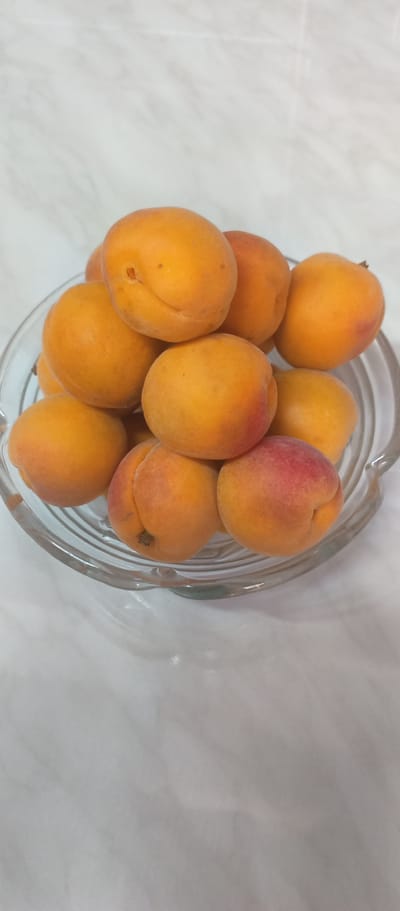 A bowl of ripe apricots sits on a marble-patterned surface. The apricots are predominantly orange with some hints of red blush. The bowl is clear glass with a ribbed design around its base. The background is a light-colored, slightly textured surface. There are no people or any other objects visible in the image. The scene appears to be a still life, likely in a domestic setting. The location in Balkanabat, Turkmenistan is not determinable from the visual information provided.
