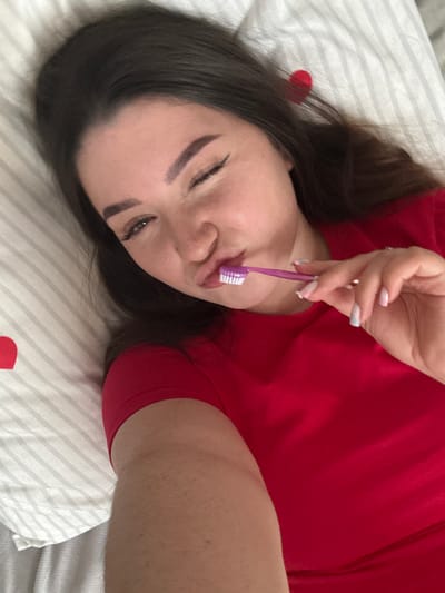 A young woman is depicted lying on a white surface with gray stripes, likely a bed or pillow, seen from a slightly overhead, tilted angle. She has long, dark brown hair framing her face and is wearing a red t-shirt. Her eyes are partially closed, and her eyebrows are distinctly shaped with makeup. She holds a purple toothbrush with white bristles to her mouth, with her lips slightly pursed around it, suggesting an action related to brushing teeth or a playful pose.

Her left hand, with neatly manicured nails painted in light, possibly iridescent or ombré shades, grips the toothbrush handle. A small, red, heart-shaped object is partially visible on the striped bedding to the left of her head. The lighting is soft and even, typical of an indoor setting. The woman's expression appears relaxed and perhaps a bit sleepy or whimsical. No specific time of day, weather, or discernible location cues are evident from the image itself, beyond being indoors. There is no visible text.