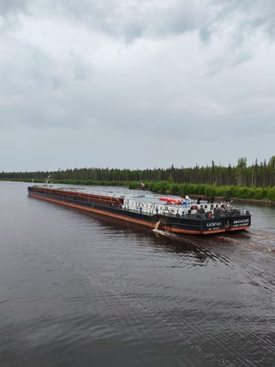 The image shows a long, dark-colored barge with an orange stripe along its hull, moving on a dark body of water. The barge is loaded with rows of what appear to be cylindrical cargo containers, colored reddish-brown. On the stern of the barge, the words "КАПИТАН" and "АФАНАСЬЕВ" are visible in white lettering, along with "САНКТ-ПЕТЕРБУРГ" and an IMO number. Several people are present on the deck of the barge. The barge is creating a wake as it moves.
In the background, a dense line of green trees and vegetation runs along the shoreline. The sky is overcast with gray clouds. The scene appears to be a canal or river, with the barge navigating through it. The location is identified as Kurdyug, Russia, suggesting this waterway is part of its geography.