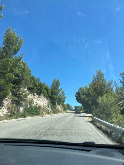 The image shows a paved road winding uphill through a landscape with rocky terrain and lush green trees on the left. The sky is a clear, bright blue with a few wispy white clouds. A silver SUV is visible driving ahead on the road. The perspective suggests the photo was taken from the driver's seat of a vehicle. The scene is characteristic of a scenic route in a Mediterranean coastal area, possibly within the Gargano National Park near Mattinata, Italy, known for its rugged coastline and abundant Mediterranean vegetation. The overall impression is one of a sunny day on a road trip through a natural, undeveloped environment.