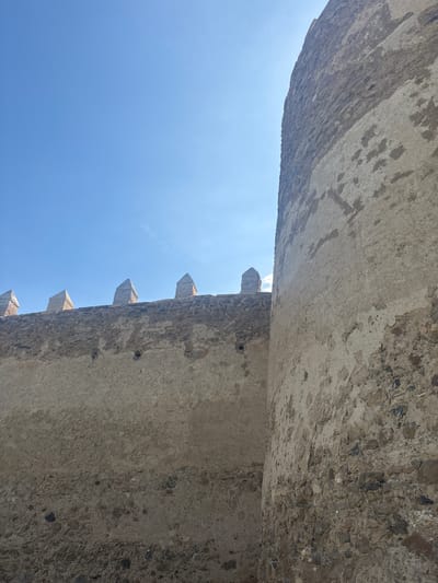 The image shows an upward view of stone walls, likely of a castle. The walls are made of gray stone with visible mortar and some weathering. The walls form a narrow gap where the blue sky is visible. One wall has crenellations, indicative of a defensive structure. The walls appear to be part of a medieval or historic building in Vera de Moncayo, Spain.