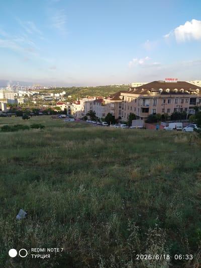 The image depicts a scenic overview of Avcılar, Turkey, on a clear day with scattered clouds. The foreground is dominated by a grassy slope with tall, dry grasses and some scattered wildflowers. In the midground, a residential area with several multi-story apartment buildings is visible. One prominent building features a dark, sloped roof with dormer windows and a sign that reads "YILDIZLAR YAPI CİHANGİR KONAKLARI." A row of vehicles, including cars and vans, are parked along the street in front of these buildings. Beyond the residential area, a port or industrial zone is visible in the distance, characterized by large cranes and storage tanks, likely part of the Istanbul port facilities. The sky is a bright blue, with the sun casting a warm light on the scene, suggesting it is either morning or late afternoon. There are no people clearly identifiable in the scene. The overall impression is one of a developing urban area with a mix of residential and industrial development against a natural backdrop.