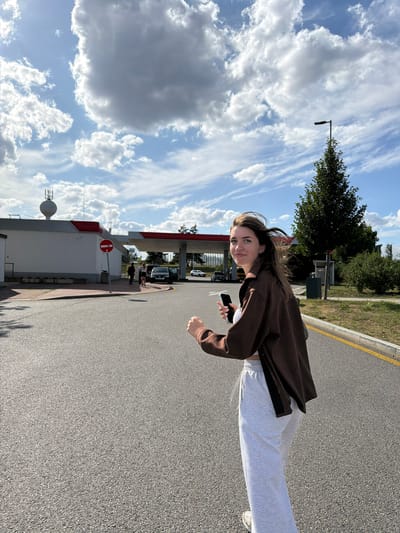 A young woman with long brown hair is looking at the camera with a slight smile. She is wearing a brown hoodie and light-colored sweatpants. She is holding a black mobile phone in her right hand. The scene is outdoors, with a paved road and a grassy area in the background. There are trees and a gas station with pumps visible. The sky is blue with scattered white clouds. The image is taken from a low angle, giving a slightly distorted perspective of the woman and the surroundings. The lighting suggests it is daytime, likely late afternoon due to the warm light. There is a "no entry" sign visible at the gas station.
